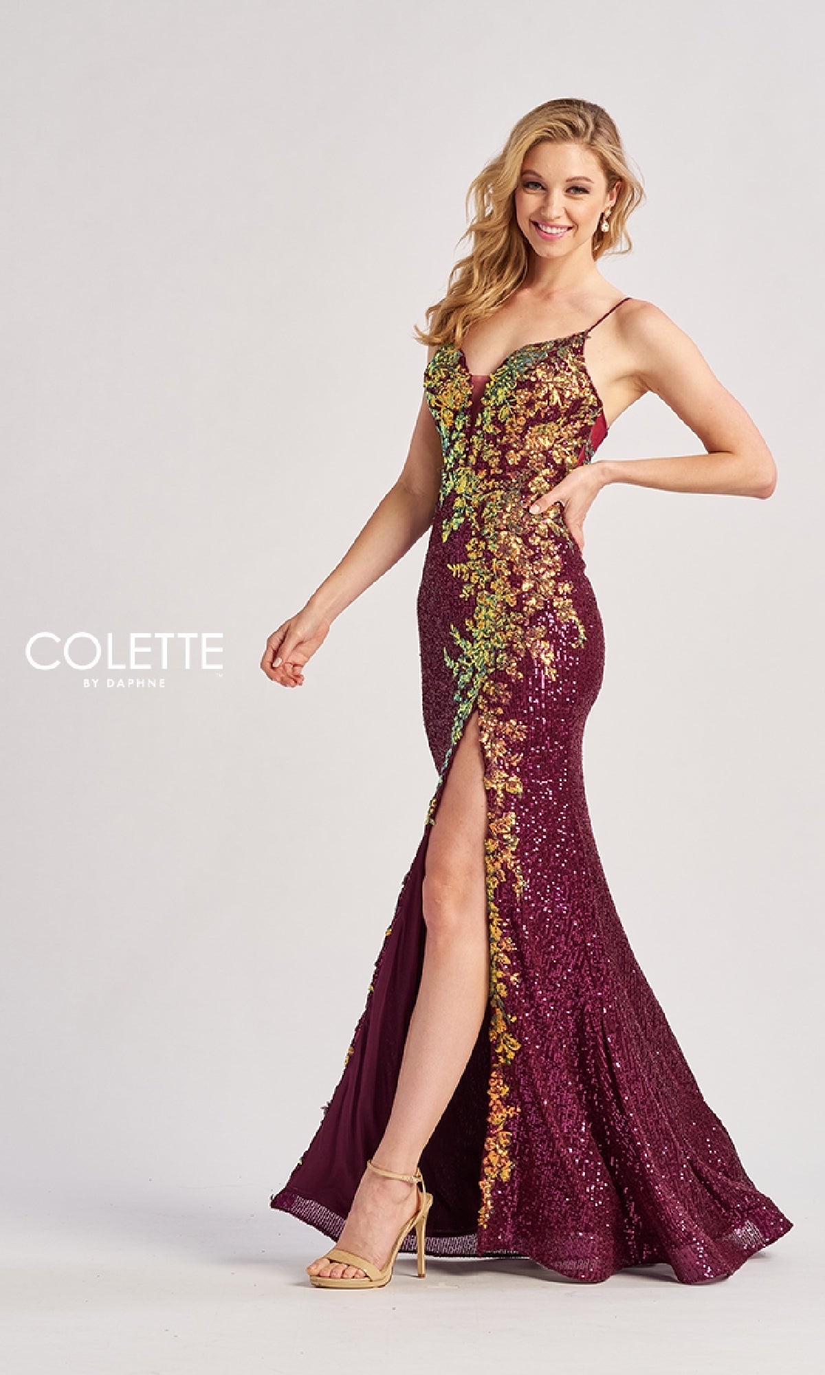 A woman poses and smiles against a plain background wearing the Colette CL8425, a sleeveless, floor-length prom dress with floral sequin appliqué, thigh-high slit, and shimmering sequins.